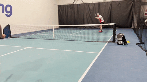 Two pickleball players practicing a pressure drill, one at the baseline and one at the net on an indoor indoor court