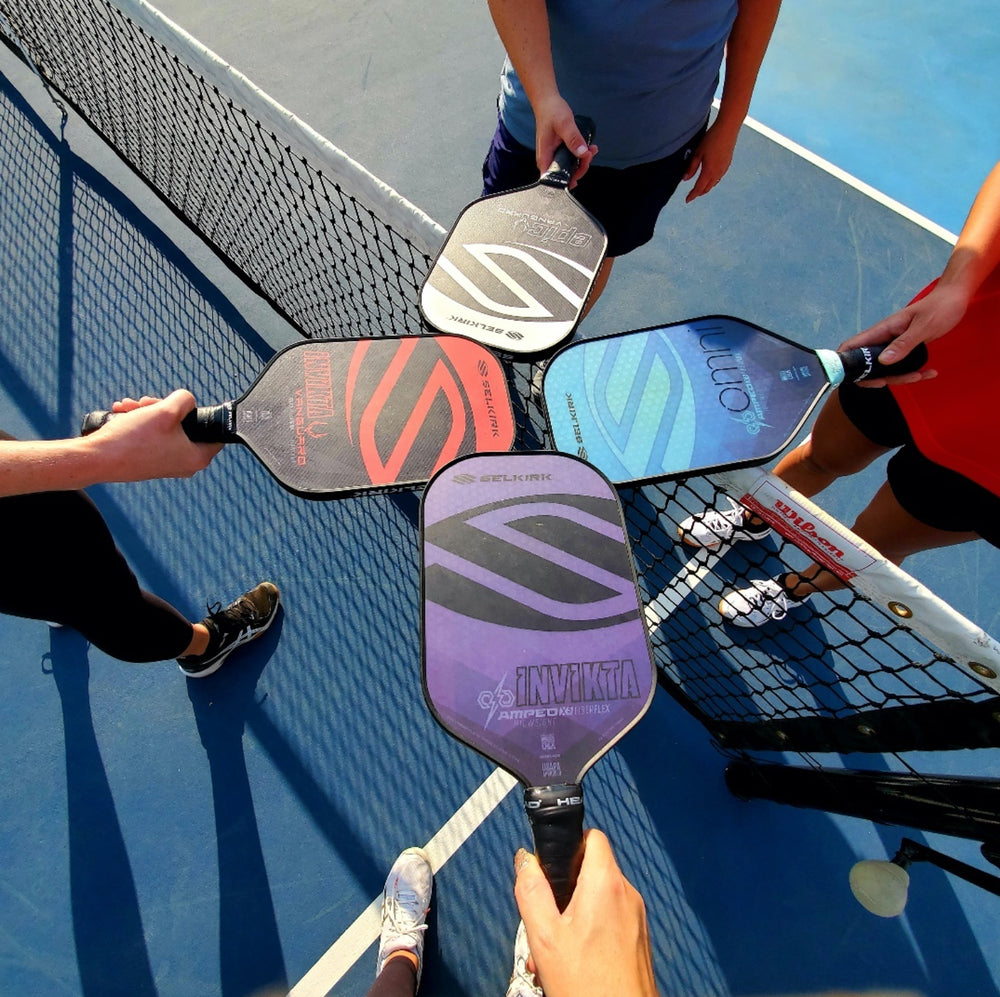 Four players touching pickleball paddles together above the net on a blue outdoor court, top-down perspective