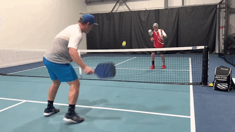 Two players trading low dinks at an indoor pickleball court near the net, one in a gray shirt and blue shorts and the other in a red jersey
