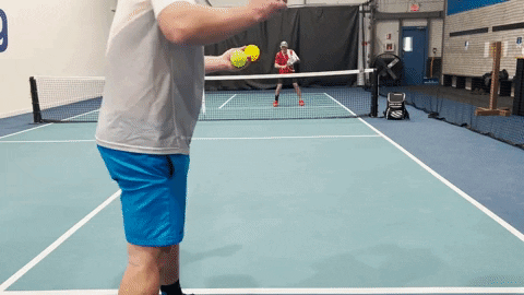 Two pickleball players on an indoor court, front player hitting a low drive while opponent prepares at the net