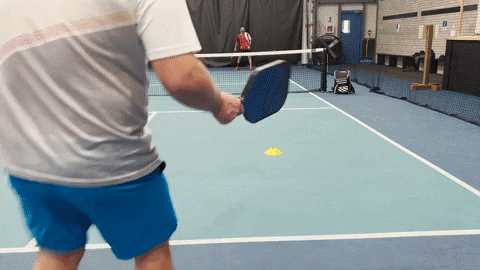 Pickleball player driving a short return crosscourt on an indoor court while an opponent waits at the baseline