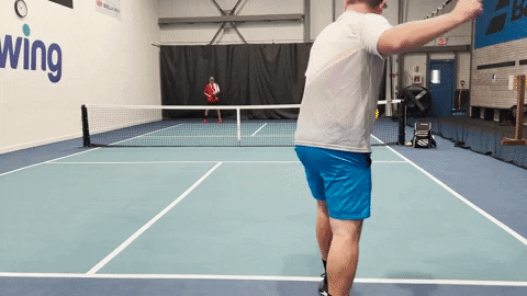 Player in blue shorts serving while partner at the baseline prepares to return on an indoor pickleball court