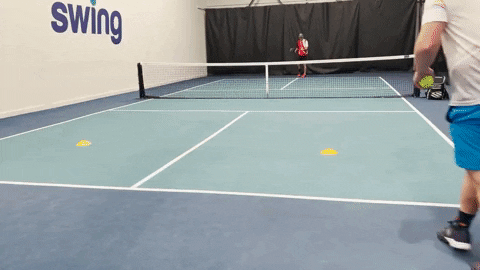 Player hitting a forehand during a singles drill on an indoor pickleball court with cones and an opponent at the baseline