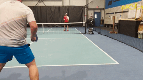 Two pickleball players practicing a drill: foreground player prepares to hit while opponent waits near the net on an indoor court