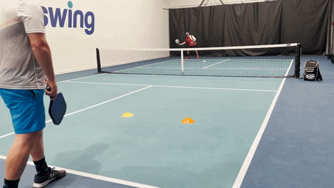 Player performing a wide-body T serve while opponent readies a return on an indoor pickleball court